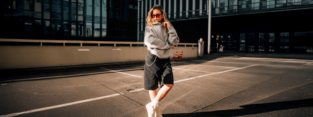 Frau trägt grauen Oversize-Hoodie und schwarze Denim Shorts im Streetwear-Look auf leerem Parkdeck – Straight Outta Cotton®