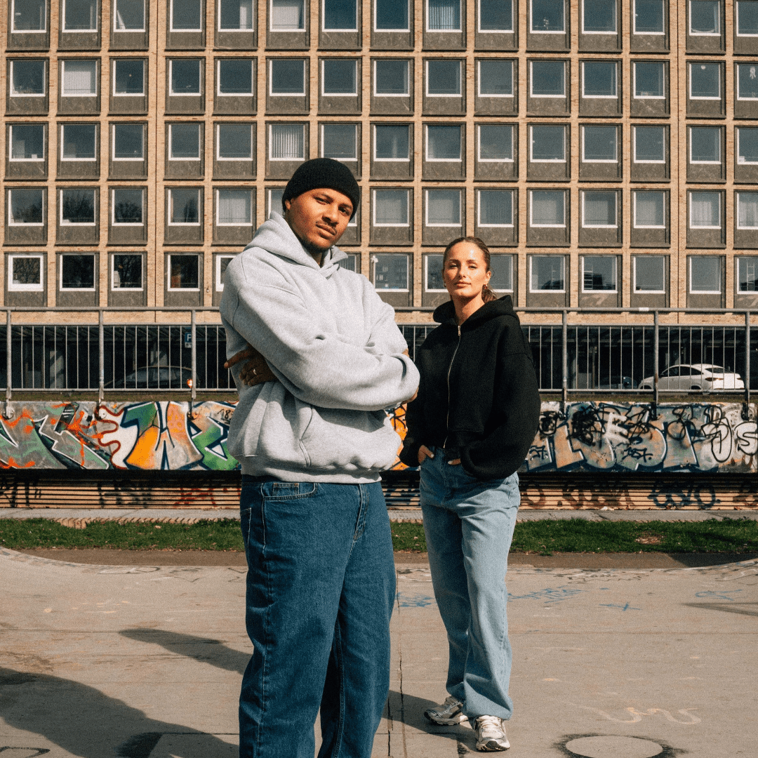 Zwei Personen im Streetwear Look mit Oversize Hoodie und Baggy Jeans vor Graffiti-Wand im Skatepark